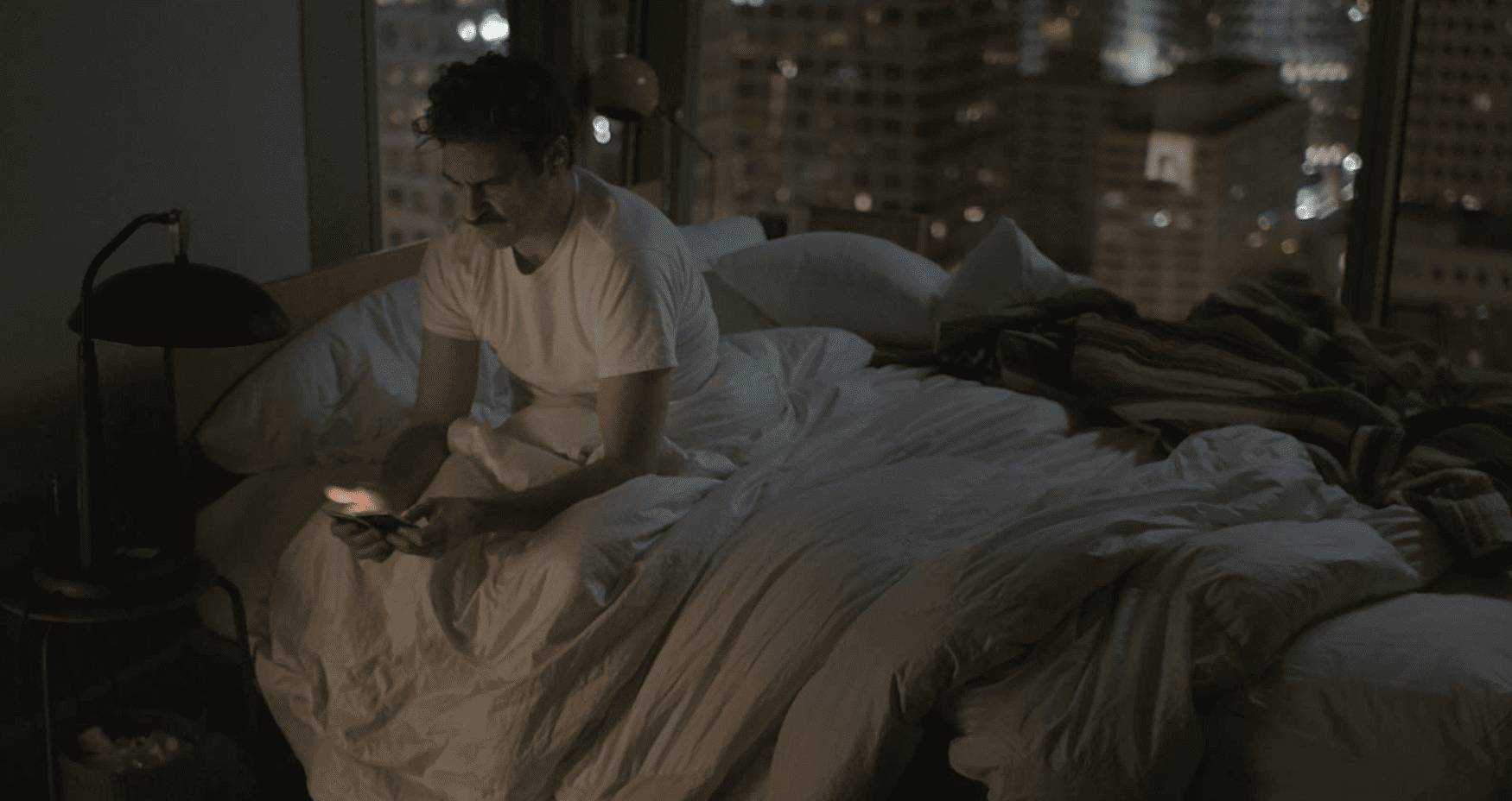 A man sits on the edge of his bed, looking at his phone in this image from Apple TV Plus