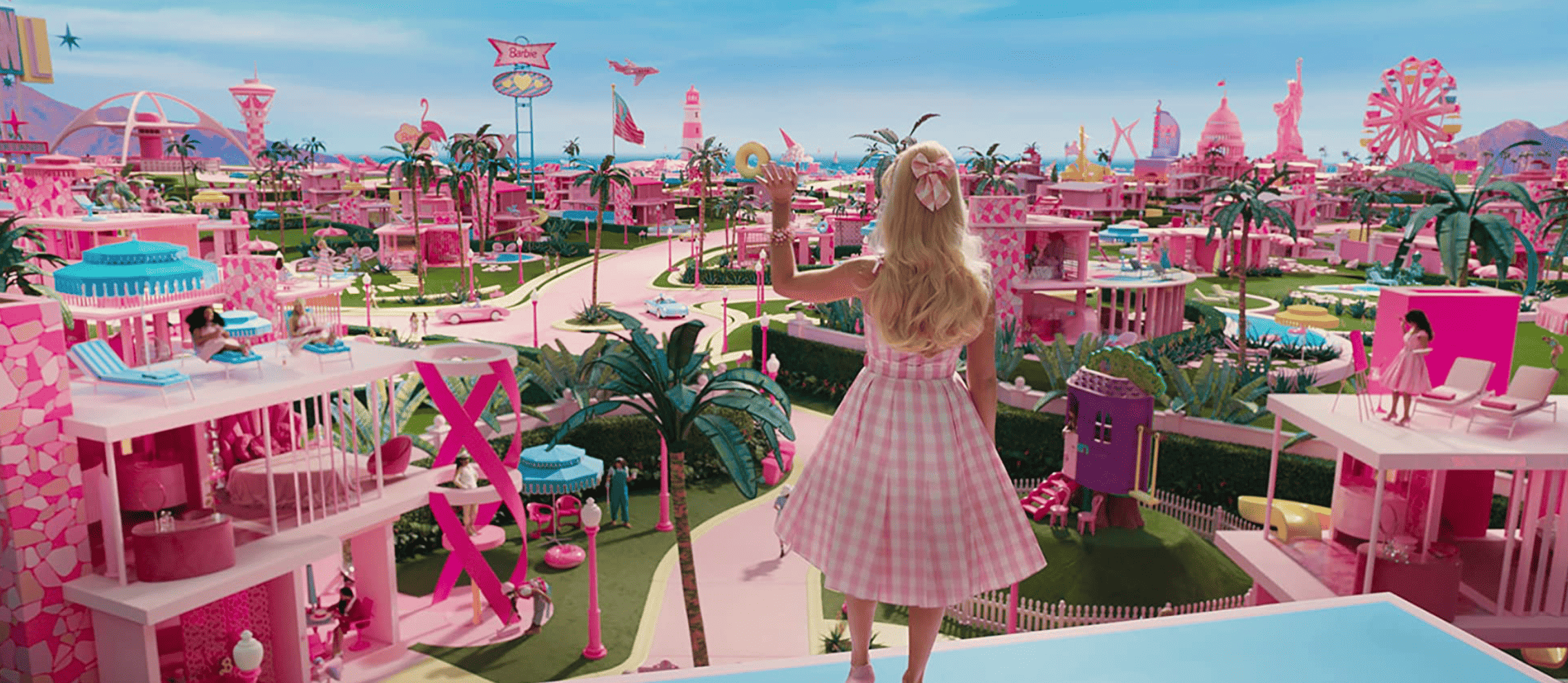 Barbie (Margot Robbie) with her back turned to the camera stands on a roof overlooking Barbie Land, a collection of doll houses and props predominantly in a bright pink color in this image from HeyDay Films.