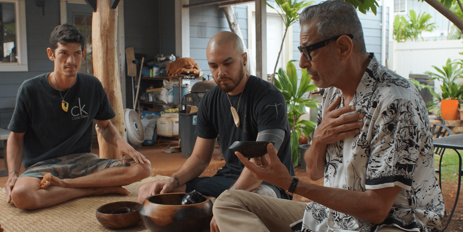 Jeff Goldblum sitting on the floor with his legs crossed while talking to locals